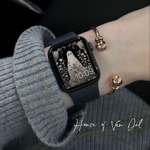 May include: A black smartwatch displaying a ghost design, with a white ghost figure and floral details. The watch is worn with a gold bracelet. The time shown is 10:09. The text "House of Van Del" is visible.