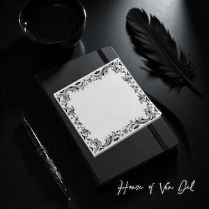 May include: A black notebook with a white square notepad featuring an ornate black and white border. A black pen and a black feather are also present. The text "House of Van Del" is written in white.