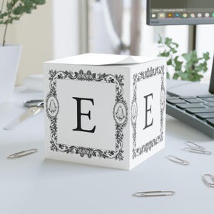 May include: White cube-shaped box with the letter "E" in black on three sides. Each side features a decorative black and white frame with floral and ghost motifs. The box sits on a white surface with scattered paper clips.