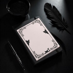 May include: A black notebook with a light pink notepad featuring a decorative black floral border and black cat illustrations. A black pen and a black feather are also visible.