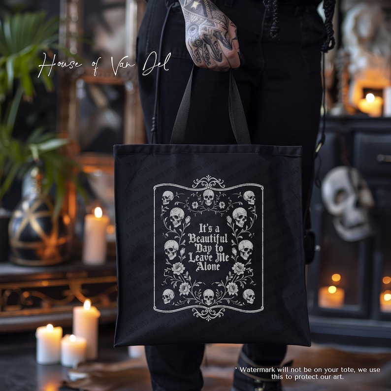 May include: Black tote bag with a white framed design featuring skulls and flowers, and the text "It's a Beautiful Day to Leave Me Alone." The bag is held by a person with tattoos. The words "House of Van Del" are written in cursive.