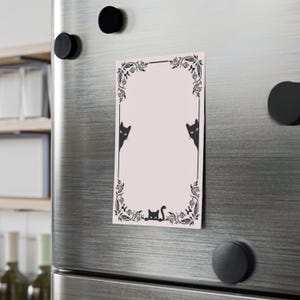 May include: A rectangular pink magnet with a black decorative border and cat illustrations. The border features floral and leaf designs. Two cats peek from the sides, and one cat is at the bottom. The magnet is attached to a silver refrigerator with black magnets.