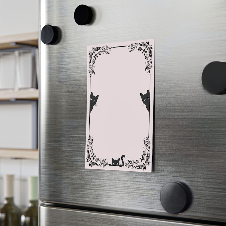 May include: A rectangular pink magnet with a black decorative border and cat illustrations. The border features floral and leaf designs. Two cats peek from the sides, and one cat is at the bottom. The magnet is attached to a silver refrigerator with black magnets.