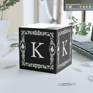 May include: Black cube-shaped notepad with the letter "K" in white. The cube features a decorative white frame with ghost and skull motifs. The notepad is on a white surface with paper clips.