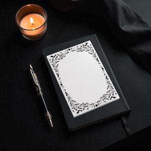 May include: A black notebook with a white notepad featuring a black floral and moon phase border. A lit candle in a glass and a decorative pen are also on the black surface.