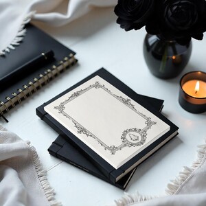 May include: A rectangular cream-colored card featuring a detailed black ornate border with intricate scrollwork and a small ghost illustration in the bottom right corner, resting on top of a black notebook with a matching black pen nearby.