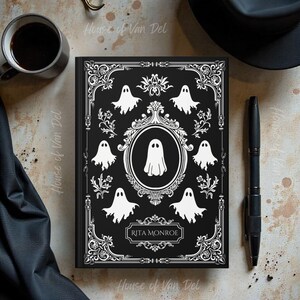 May include: Black journal with a white ghost design. The cover features a central oval with a ghost and is surrounded by smaller ghosts and decorative flourishes. The name "Rita Monroe" is printed at the bottom. A black pen and coffee cup are visible.