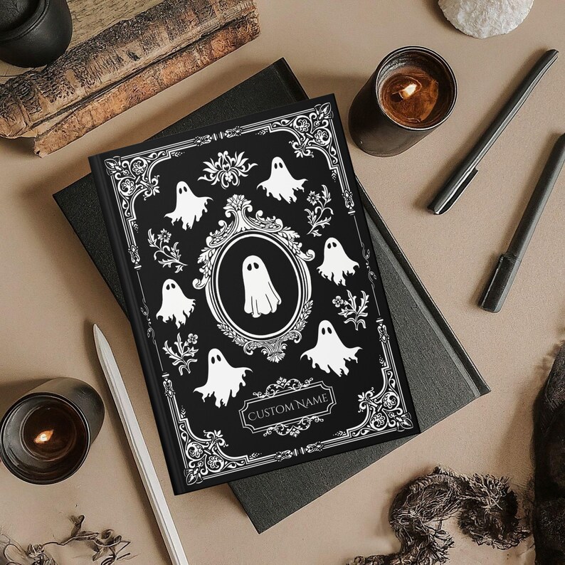 May include: A black journal with a white ghost design and the words "CUSTOM NAME". The cover features a central oval with a ghost illustration, surrounded by smaller ghost figures and decorative floral elements. Candles and pens are also visible.