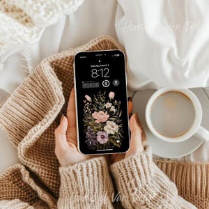 May include: A smartphone with a floral wallpaper and a digital clock displaying "Monday, March 3 8:12" on the screen. The phone is being held by a person wearing a brown knitted jumper.