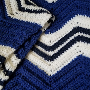 May include: A close-up of a blue and white crocheted blanket with a chevron pattern. The blanket is made with thick yarn and has a soft, textured feel.