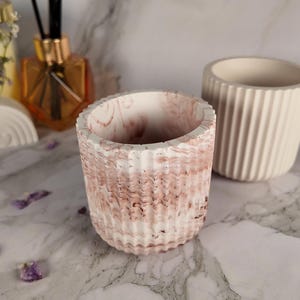 Op de afbeelding: Twee decoratieve betonnen plantenbakken. Eén heeft een getextureerd, roze en wit gemarmerd ontwerp. De andere is een effen, gebroken witte plantenbak met verticale ribbels. De plantenbakken staan op een marmeren oppervlak.