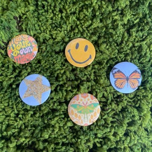 May include: Four circular pinback buttons featuring distinct designs: a multicolored graphic with the text 'make your own magic', a yellow smiley face, a light blue button with a gold glitter star, and a moth illustration on a floral background.