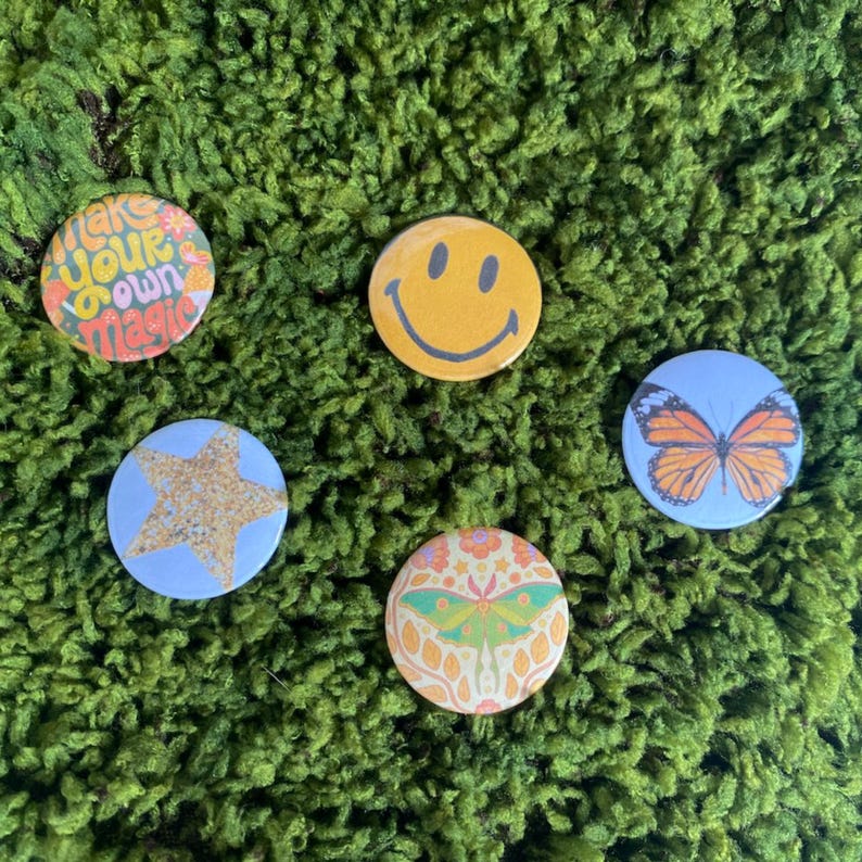 May include: Four circular pinback buttons featuring distinct designs: a multicolored graphic with the text 'make your own magic', a yellow smiley face, a light blue button with a gold glitter star, and a moth illustration on a floral background.