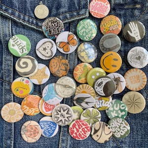 May include: A collection of circular pin-back badges featuring diverse graphic designs including a butterfly, a compass, a hand, a smiley face, a clover, and various text phrases like Stay Wild Moon Child, Stay Kind, and This Is My Happy Place.