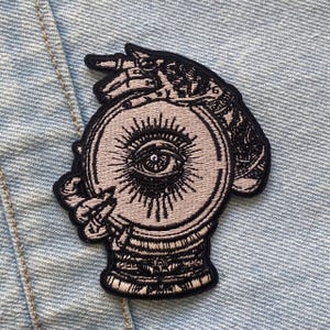 May include: Embroidered patch depicting a crystal ball with an eye design, held by a skeletal hand. The patch has a black border and a light grey and black colour scheme. Suitable for clothing or accessories.