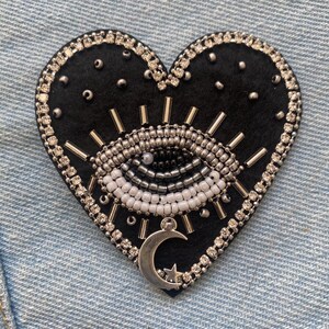 May include: A heart-shaped patch with a black base, adorned with a beaded eye design in white, silver, and black. The patch is edged with silver rhinestones and includes a silver crescent moon and star charm.