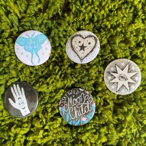 May include: Five circular pinback buttons featuring various designs: a light blue moth, a black and white heart with a star, a black and white starburst with an eye, a black hand with symbols, and a dark button with the text Stay Wild Moon Child.
