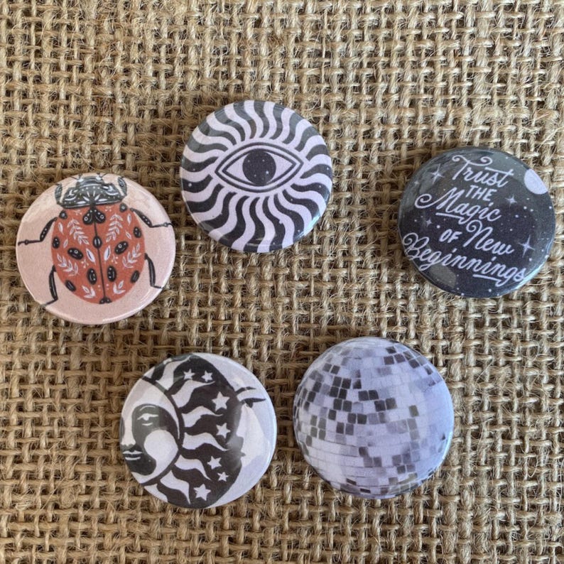 May include: Five circular pinback buttons featuring distinct designs: a ladybug with floral patterns, a black and white sunburst eye, a dark circle with the text Trust the Magic of New Beginnings, a crescent moon face, and a silver disco ball pattern.