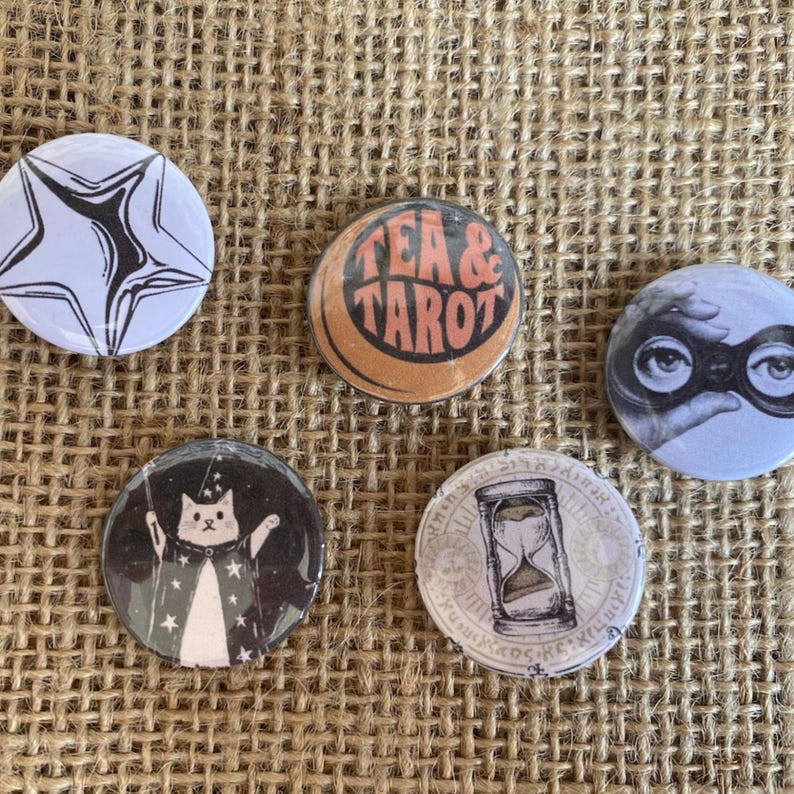 May include: Five circular pinback buttons featuring distinct designs: a black and white star, an orange circle with the text TEA & TAROT, a black and white illustration of eyes looking through binoculars, a cat in a star-patterned robe, and an hourglass.
