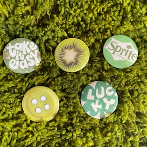 May include: A set of five circular pinback buttons featuring various green designs: one with the text 'take it easy' and small hearts, a realistic kiwi fruit slice, a Sprite logo, a yellow four-hole button graphic, and a clover with the text 'LUCKY'.