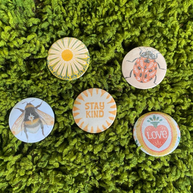 May include: Five circular pinback buttons featuring distinct designs: a yellow and white daisy, an orange ladybug with black spots, a bumblebee, a sunburst graphic with the text STAY KIND, and a heart-shaped emblem with the word LOVE.