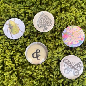 May include: Five circular pin-back buttons featuring distinct designs: a yellow beetle, a grey spade symbol, a colorful floral graphic with the text DREAM BIG, a black ampersand symbol on a beige background, and a grey butterfly illustration.