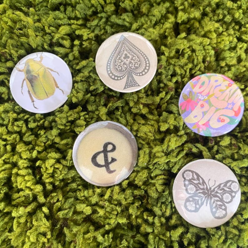 May include: Five circular pin-back buttons featuring distinct designs: a yellow beetle, a grey spade symbol, a colorful floral graphic with the text DREAM BIG, a black ampersand symbol on a beige background, and a grey butterfly illustration.
