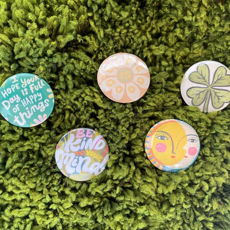May include: Four circular pinback buttons with distinct designs. One is teal with the text "I hope your day is full of happy things". One is a peach flower with "Life is good" text. One is a green four-leaf clover. One is a yellow sun face.