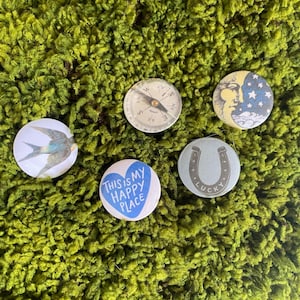 May include: A collection of five circular pin-back buttons featuring diverse designs: a blue bird in flight, a vintage-style compass, a crescent moon with stars, a blue heart with the text THIS IS MY HAPPY PLACE, and a horseshoe with the word LUCKY.