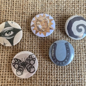 May include: Five circular pin-back buttons featuring distinct designs: a black and white eye, a golden sun with a face, a black and white spiral, a black and white butterfly with floral patterns, and a blue horseshoe with the word LUCKY.
