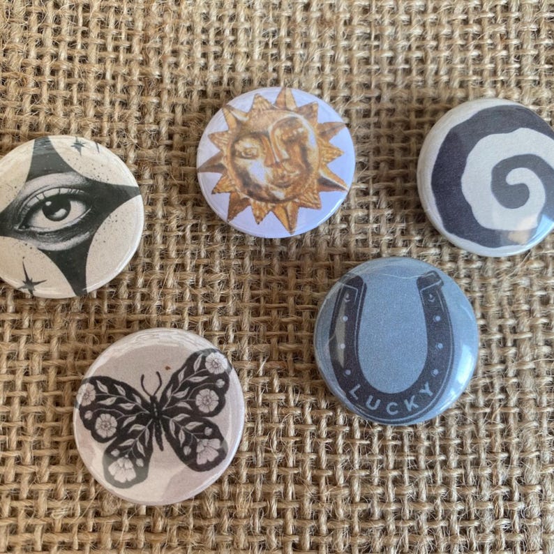 May include: Five circular pin-back buttons featuring distinct designs: a black and white eye, a golden sun with a face, a black and white spiral, a black and white butterfly with floral patterns, and a blue horseshoe with the word LUCKY.