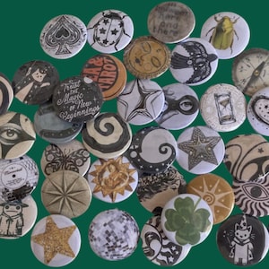 May include: A collection of circular pin-back badges featuring diverse graphic designs including celestial motifs, insects, tarot-inspired imagery, stars, eyes, a compass, a four-leaf clover, and text such as Trust the Magic of New Beginnings.