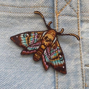 May include: Embroidered moth patch with detailed stitching. The patch has a brown body, wings with teal, pink, and gold accents, and fine antennae. The patch is set against a light blue denim background.