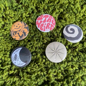 May include: Five circular pinback buttons featuring diverse designs: an orange pumpkin character, a pink button with the text "STAY WEIRD", a black and white spiral, a black crescent moon with botanical accents, and a silver starburst pattern.