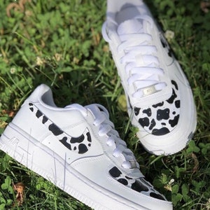 May include: White leather trainers with a black and white cow print design. The trainers have a white swoosh logo on the side and a black and white cow print design on the toe.