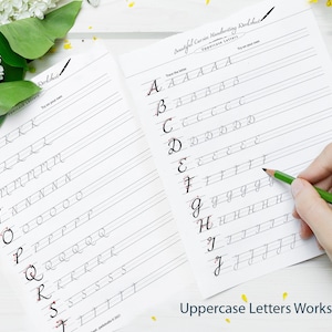 May include: Two pages of a cursive handwriting worksheet for uppercase letters. The worksheet has lines for tracing the letters. A hand is holding a green pencil and tracing the letter 'J'.