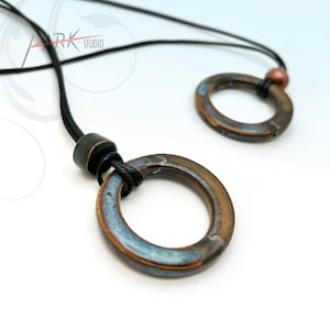May include: A necklace with two ceramic rings, one larger than the other, both with a blue and brown glaze. The necklace is made with black cord and has a copper bead.