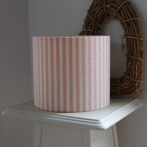 May include: A drum lampshade featuring a pink and white striped design. The lampshade is placed on a white table. A brown twig wreath is visible in the background.