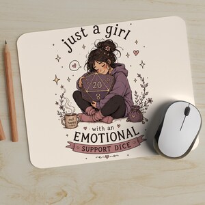 May include: A rectangular mousepad featuring a cartoon illustration of a person hugging a large purple twenty-sided die. The text reads just a girl with an EMOTIONAL SUPPORT DICE. A small mug labeled roll happy and a pouch are nearby, surrounded by stars and hearts.