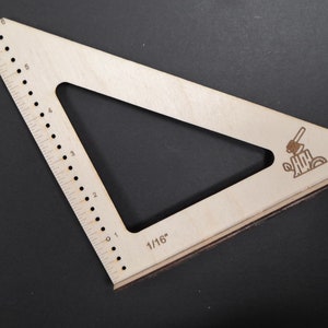 May include: A wooden triangle ruler with a 1/16 inch scale and a small axe and log design. The ruler has a 6-inch scale on one side.
