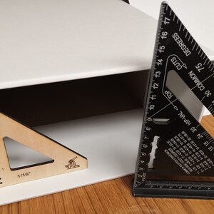 May include: A white box with a wooden triangle ruler inside. The ruler has markings in 1/16 inches. A black metal triangle ruler is partially visible on the right side of the image. The ruler has markings in degrees and inches.
