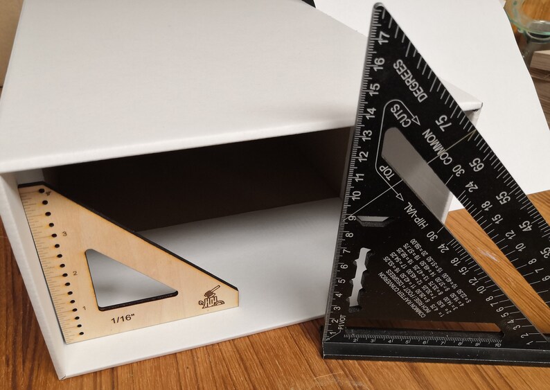 May include: A white box with a wooden triangle ruler inside. The ruler has markings in 1/16 inches. A black metal triangle ruler is partially visible on the right side of the image. The ruler has markings in degrees and inches.