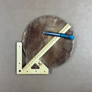 May include: A wooden protractor with a ruler and a triangle shape. The protractor is on a brown wooden surface with a craft knife.