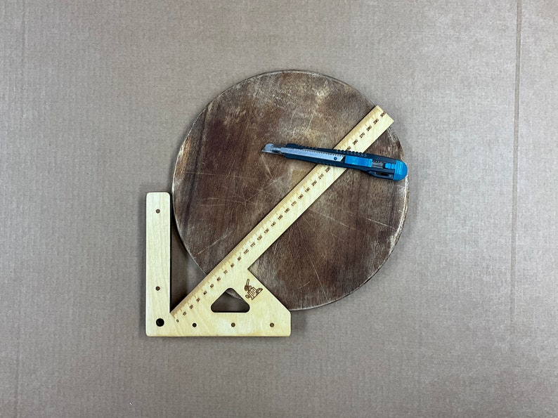 May include: A wooden protractor with a ruler and a triangle shape. The protractor is on a brown wooden surface with a craft knife.