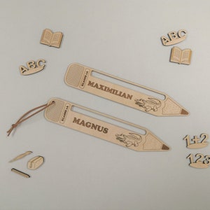 Könnte beinhalten: Zwei Holz-Lesezeichen in Bleistiftform mit einem Krokodil-Design. Die Lesezeichen sind mit den Namen "Magnus" und "Maximillian" personalisiert. Die Lesezeichen sind aus hellem braunem Holz gefertigt und haben eine Schnur am oberen Ende.