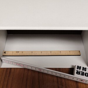 May include: A white box with a white interior. A wooden ruler with black markings is placed inside the box. A second ruler is placed outside the box, partially visible. The ruler is marked in inches and centimetres.