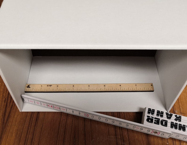 May include: A white box with a white interior. A wooden ruler with black markings is placed inside the box. A second ruler is placed outside the box, partially visible. The ruler is marked in inches and centimetres.