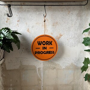 Könnte beinhalten: Runder, orangefarbener Aufhänger mit schwarzem Text "WORK - IN - PROGRESS". Das Schild hängt in einem Badezimmer über einer Toilette. Der Hintergrund ist eine strukturierte, cremefarbene Wand.