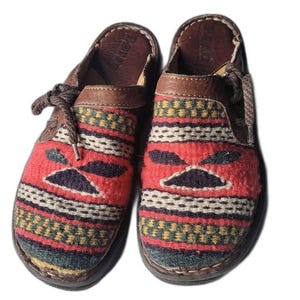 May include: Pair of slip-on shoes with a woven textile design in red, black, white, and green. The shoes have brown leather accents, laces, and soles. The textile pattern features a geometric design.