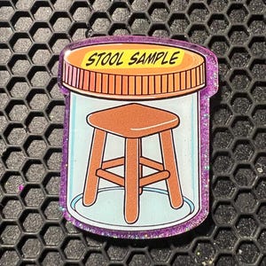 May include: A novelty pin featuring a cartoon stool inside a jar, labeled "STOOL SAMPLE" in bold text. The jar has a purple glitter border and a brown lid. The stool is brown with four legs.